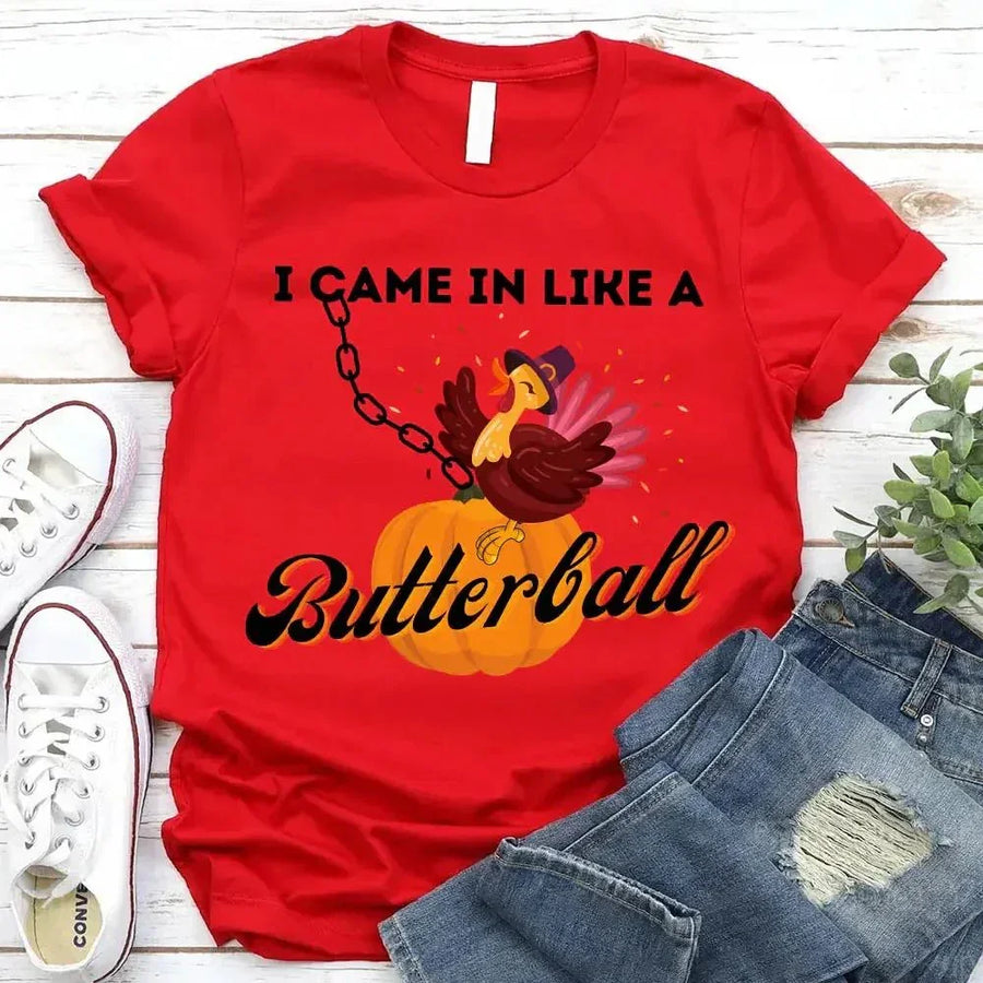 May include a casual crew-neck T-shirt with a playful Thanksgiving graphic showing a turkey stepping onto or perched on a pumpkin, surrounded by leaves or autumn motifs, with bold front text reading “I Came In Like A Butterball.”