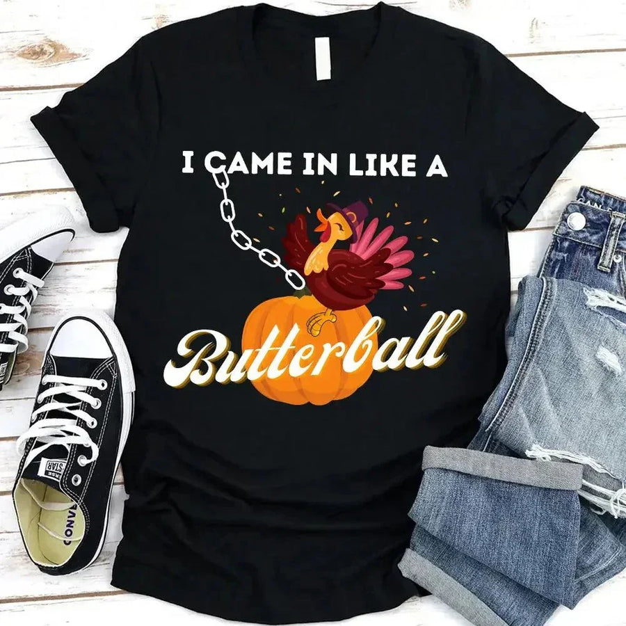 May include a casual crew-neck T-shirt with a playful Thanksgiving graphic showing a turkey stepping onto or perched on a pumpkin, surrounded by leaves or autumn motifs, with bold front text reading “I Came In Like A Butterball.”
