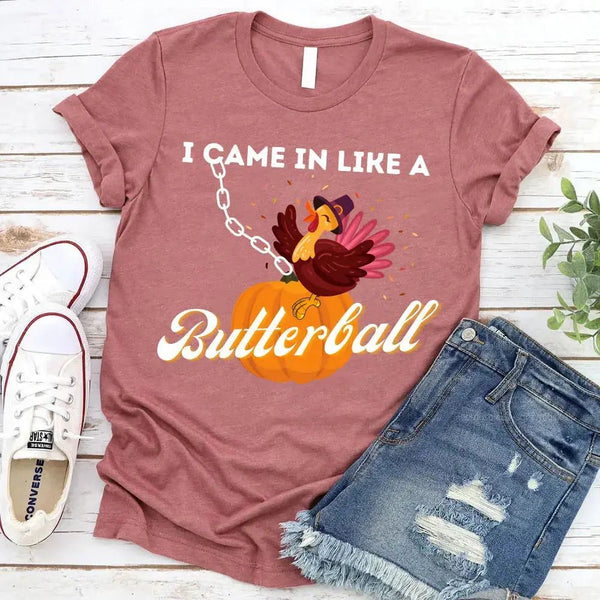 May include a casual crew-neck T-shirt with a playful Thanksgiving graphic showing a turkey stepping onto or perched on a pumpkin, surrounded by leaves or autumn motifs, with bold front text reading “I Came In Like A Butterball.”