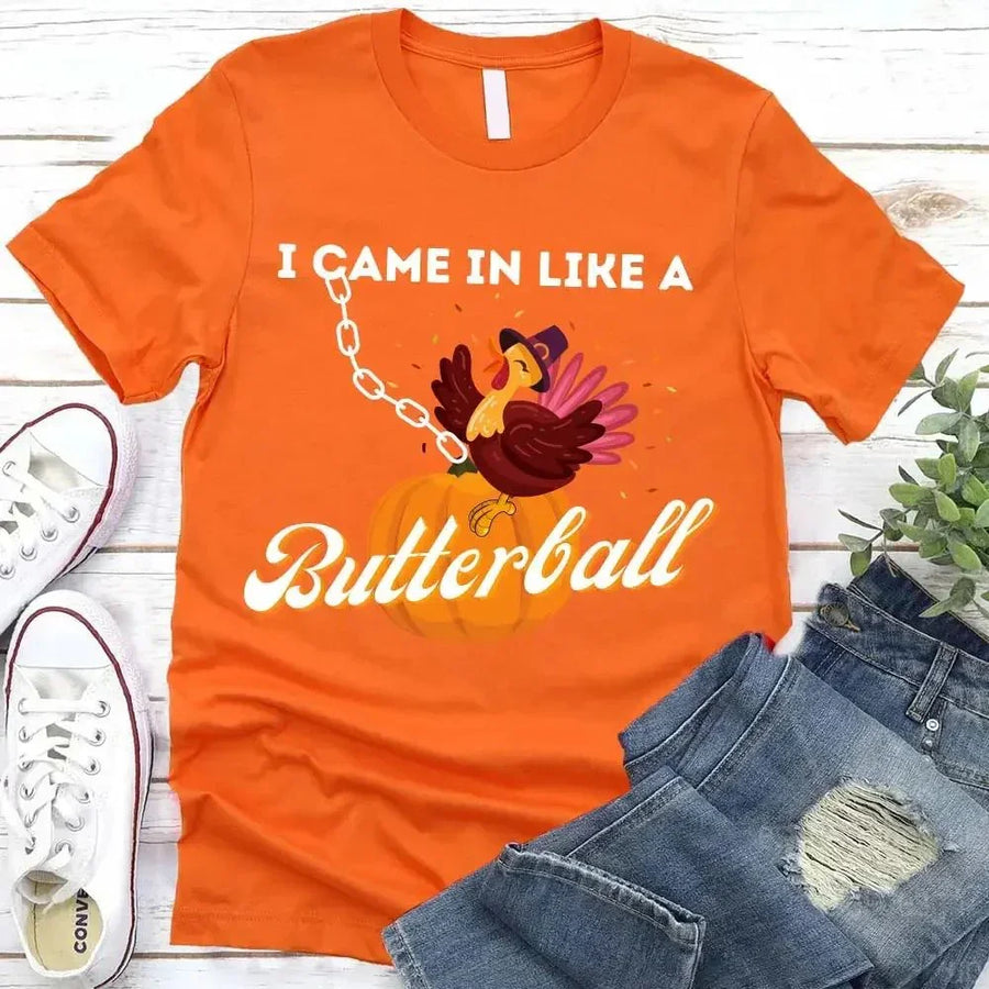 May include a casual crew-neck T-shirt with a playful Thanksgiving graphic showing a turkey stepping onto or perched on a pumpkin, surrounded by leaves or autumn motifs, with bold front text reading “I Came In Like A Butterball.”