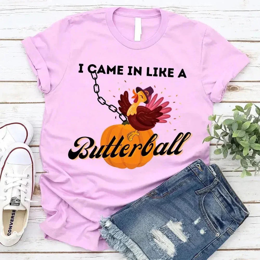 May include a casual crew-neck T-shirt with a playful Thanksgiving graphic showing a turkey stepping onto or perched on a pumpkin, surrounded by leaves or autumn motifs, with bold front text reading “I Came In Like A Butterball.”