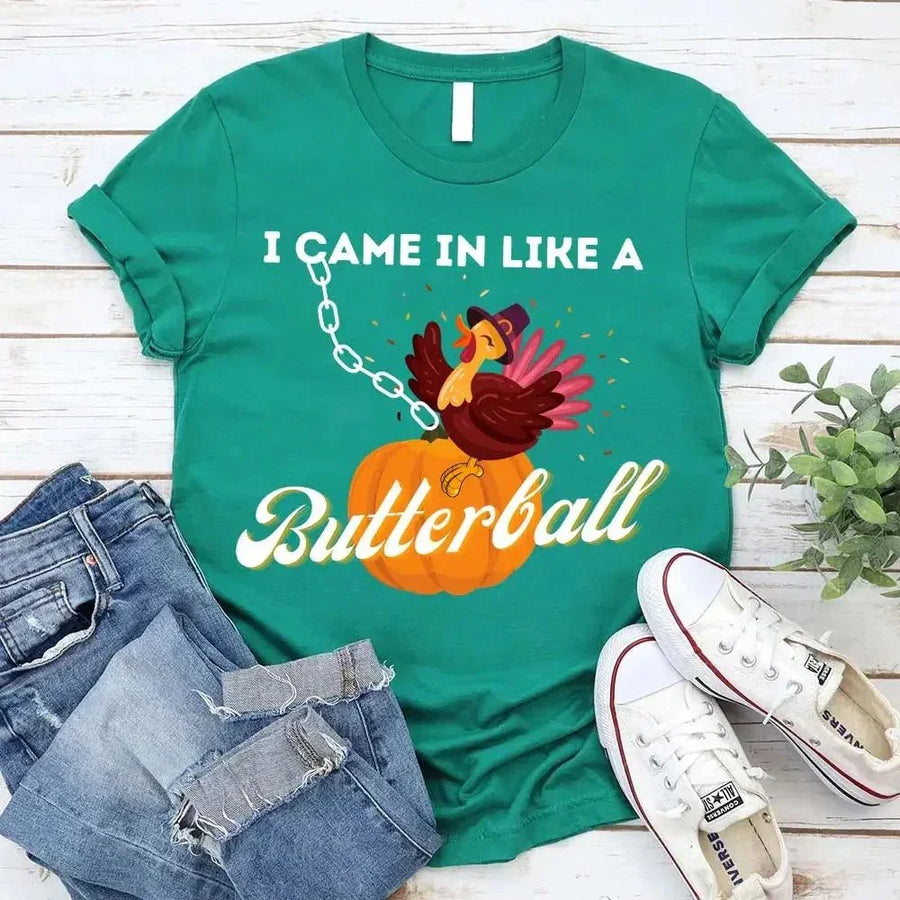 May include a casual crew-neck T-shirt with a playful Thanksgiving graphic showing a turkey stepping onto or perched on a pumpkin, surrounded by leaves or autumn motifs, with bold front text reading “I Came In Like A Butterball.”