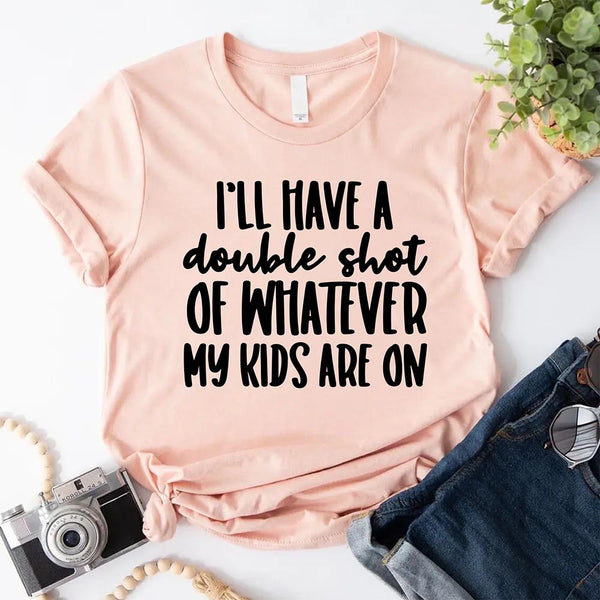 Peach t-shirt with humorous text on I'll Have a Double Shot of Whatever My Kids Are On a white surface with accessories.