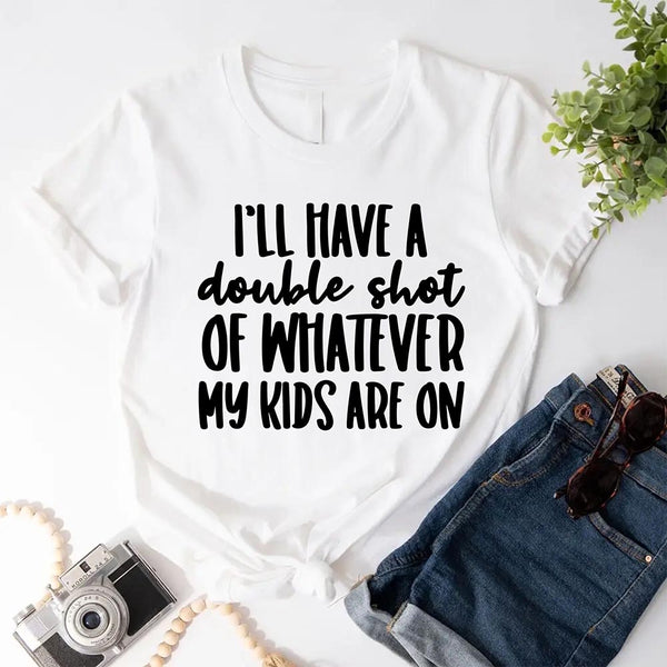 White t-shirt with text 'I'll have a double shot of whatever my kids are on' on a white background with jeans, camera, and sunglasses.