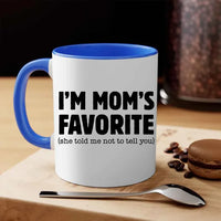 White mug with blue handle and text 'I'm Mom's Favorite (she told me not to tell you)' on a wooden surface with coffee beans and a macaron.