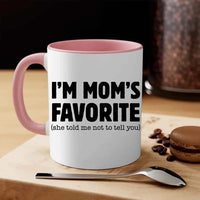 White mug with pink interior and handle, featuring text 'I'm Mom's Favorite (she told me not to tell you)' on a wooden surface with coffee beans and a macaron.
