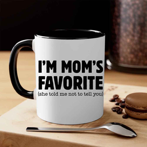 White mug with black text 'I'M MOM'S FAVORITE (she told me not to tell you)' on a wooden surface with coffee beans and a macaron.