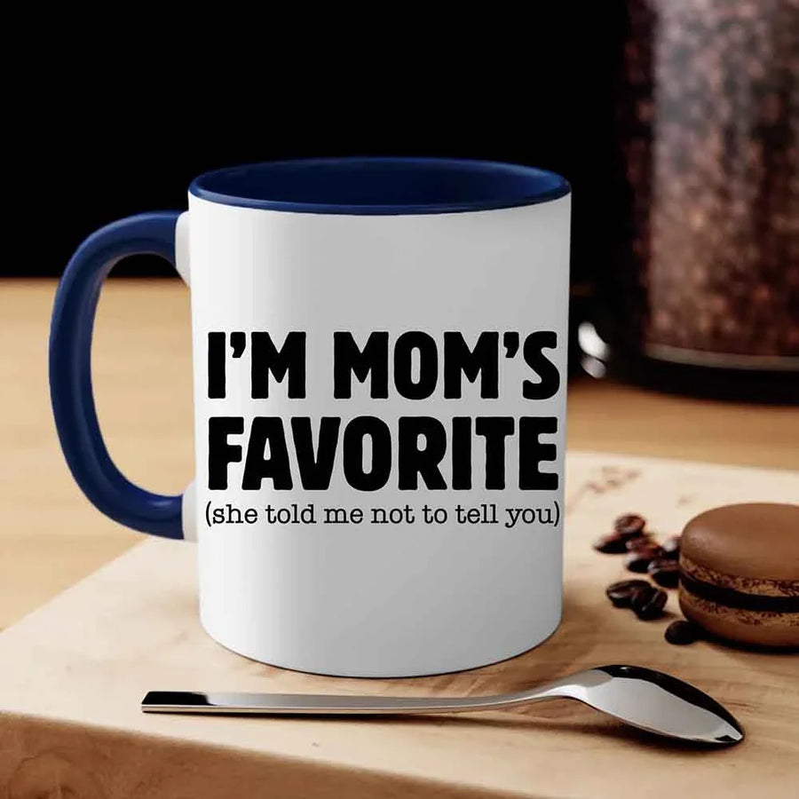 White mug with blue interior and handle, featuring text 'I'm Mom's Favorite (she told me not to tell you)' on a wooden surface with coffee beans and a macaron.