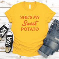 Yellow t-shirt with 'SHE'S MY Sweet POTATO' text, paired with black sneakers and blue jeans on a wooden surface.