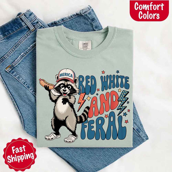 May include a folded bay Comfort Colors t-shirt with a playful raccoon graphic and bold "Red White and Feral" 4th of July text, paired with denim.