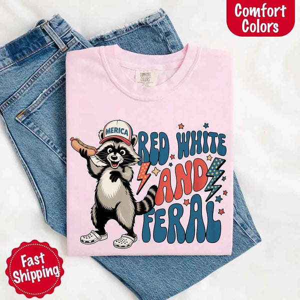 May include a folded blossom Comfort Colors t-shirt with a playful raccoon graphic and bold "Red White and Feral" 4th of July text, paired with denim.