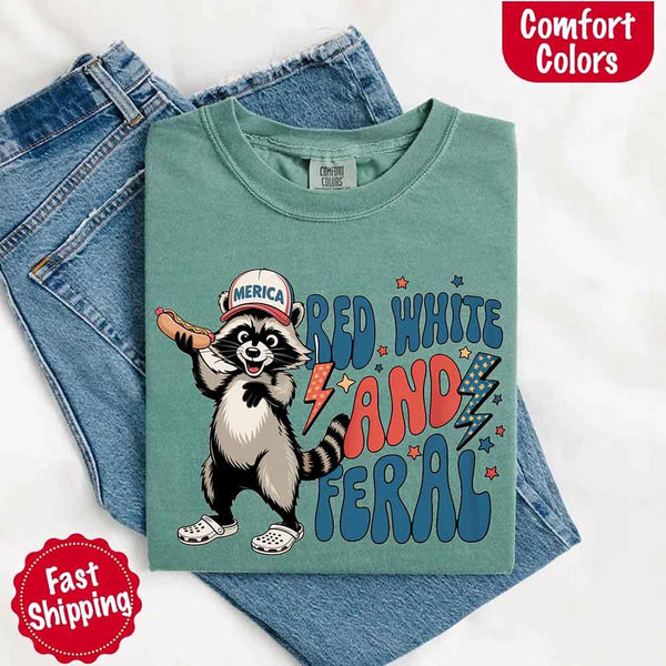 May include a folded light green Comfort Colors t-shirt with a playful raccoon graphic and bold "Red White and Feral" 4th of July text, paired with denim.