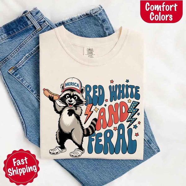 May include a folded ivory Comfort Colors t-shirt with a playful raccoon graphic and bold "Red White and Feral" 4th of July text, paired with denim.