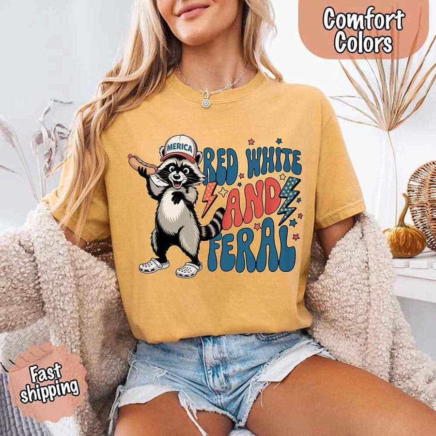 May include a woman wearing a Comfort Colors t-shirt in mustardwith a colorful "Red White and Feral" raccoon hotdog print – fun and patriotic summer design.