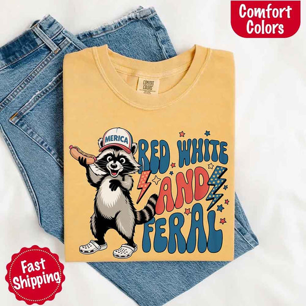 May include a folded mustard Comfort Colors t-shirt with a playful raccoon graphic and bold "Red White and Feral" 4th of July text, paired with denim.