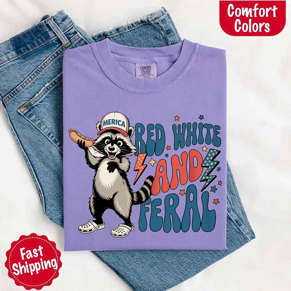 May include a folded violet Comfort Colors t-shirt with a playful raccoon graphic and bold "Red White and Feral" 4th of July text, paired with denim.