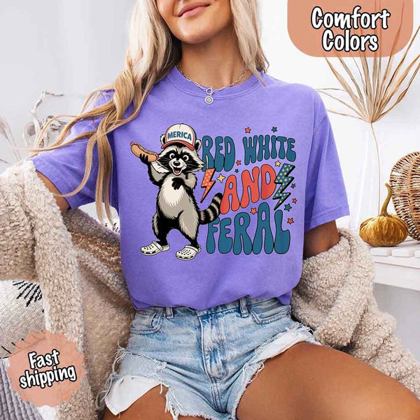 May include a woman wearing a Comfort Colors t-shirt in violet with a colorful "Red White and Feral" raccoon hotdog print – fun and patriotic summer design.