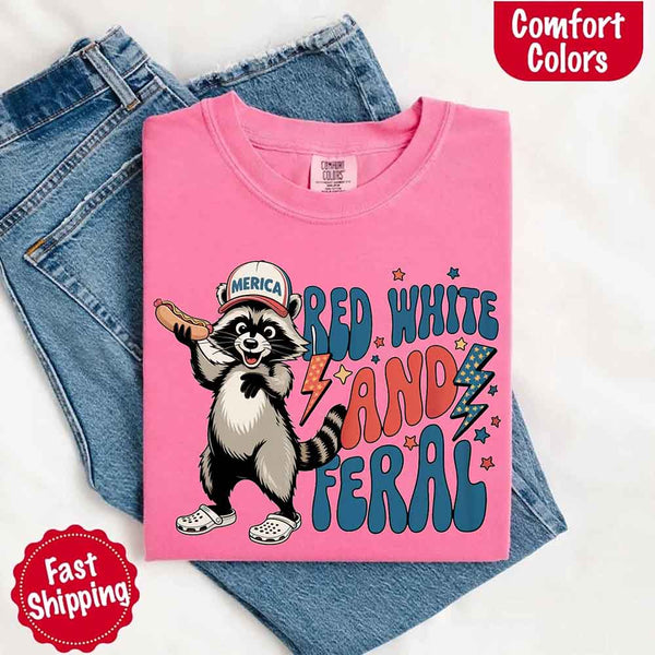 May include a folded watermelon Comfort Colors t-shirt with a playful raccoon graphic and bold "Red White and Feral" 4th of July text, paired with denim.