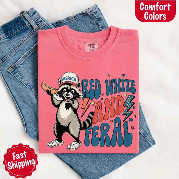 May include a folded crunchberry Comfort Colors t-shirt with a playful raccoon graphic and bold "Red White and Feral" 4th of July text, paired with denim.