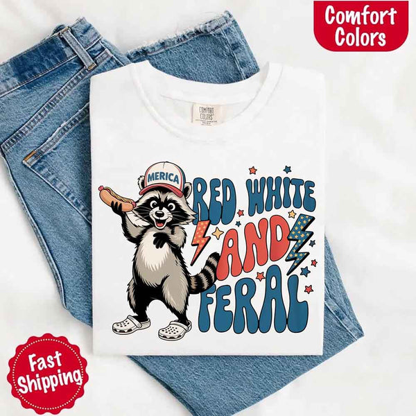 May include a folded white Comfort Colors t-shirt with a playful raccoon graphic and bold "Red White and Feral" 4th of July text, paired with denim.