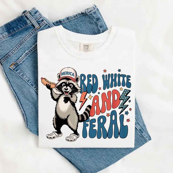 May include a folded white Comfort Colors t-shirt with a playful raccoon graphic and bold "Red White and Feral" 4th of July text, paired with denim.