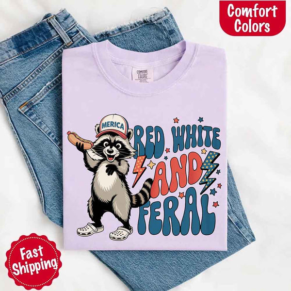 May include a folded orchid Comfort Colors t-shirt with a playful raccoon graphic and bold "Red White and Feral" 4th of July text, paired with denim.