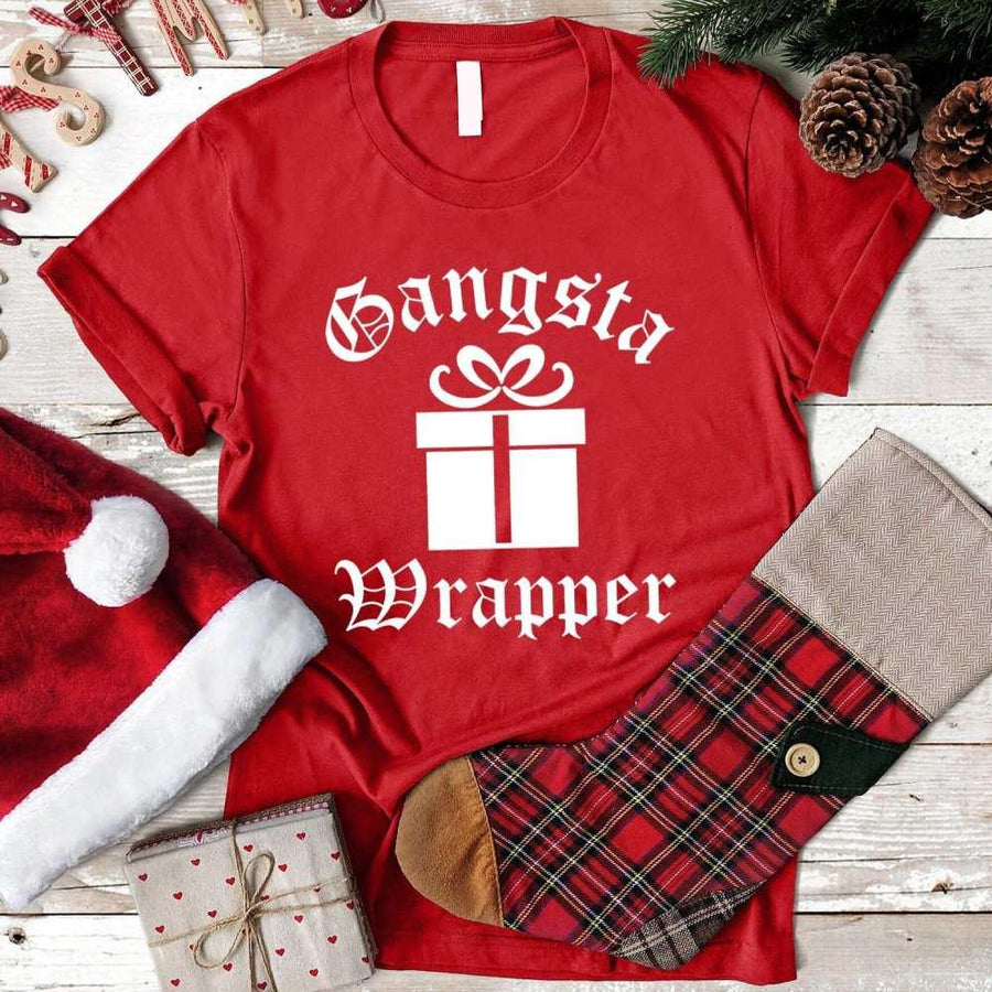 Red t-shirt with 'Gangsta Wrapper' design, Santa hat, plaid pants, and Christmas decor on a wooden surface.