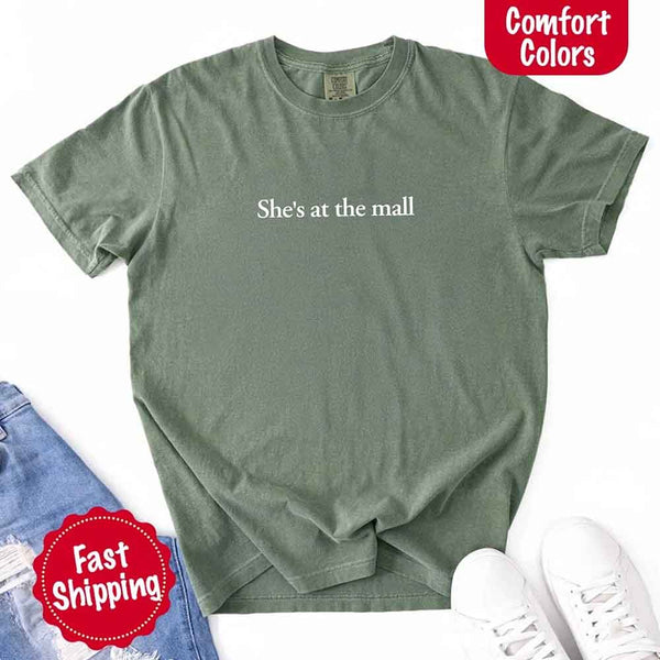 May include a light green Comfort Colors T-shirt laid flat with the phrase "She's at the mall" printed in white, styled alongside denim jeans and white sneakers, featuring fast shipping branding.