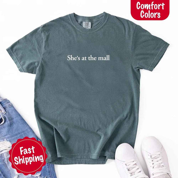 May include a denim Comfort Colors T-shirt laid flat with the phrase "She's at the mall" printed in white, styled alongside denim jeans and white sneakers, featuring fast shipping branding.