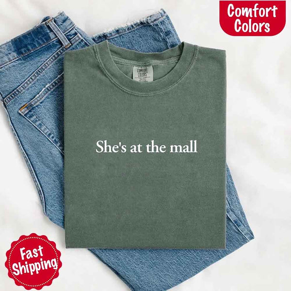 May include a folded moss Comfort Colors T-shirt with the text "She's at the mall" printed in white, styled with denim jeans and white sneakers, promoting fast shipping.