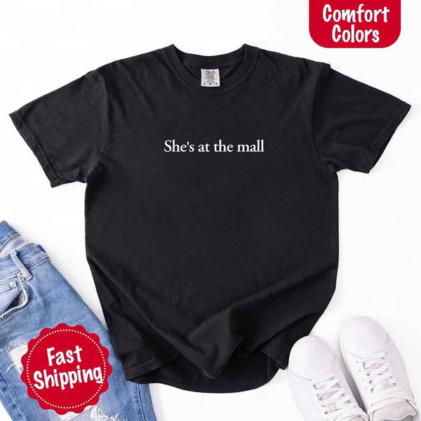 May include a black Comfort Colors T-shirt laid flat with the phrase "She's at the mall" printed in white, styled alongside denim jeans and white sneakers, featuring fast shipping branding