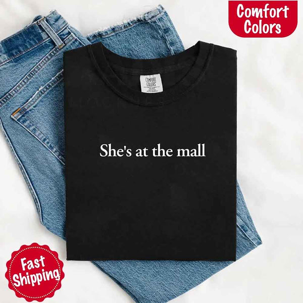 May include a folded black Comfort Colors T-shirt with the text "She's at the mall" printed in white, styled with denim jeans and white sneakers, promoting fast shipping.