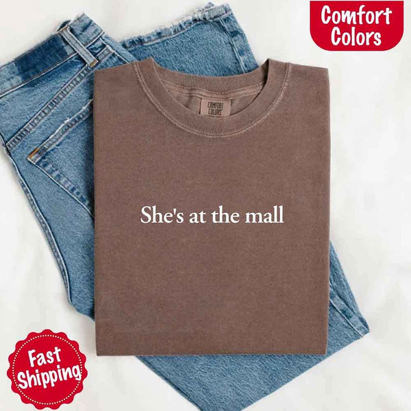 May include a folded espresso Comfort Colors T-shirt with the text "She's at the mall" printed in white, styled with denim jeans and white sneakers, promoting fast shipping.