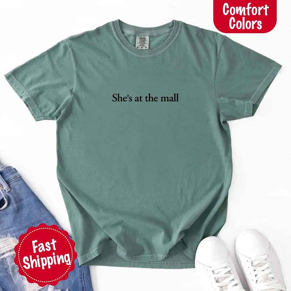May include a light green Comfort Colors T-shirt laid flat with the phrase "She's at the mall" printed in white, styled alongside denim jeans and white sneakers, featuring fast shipping branding.