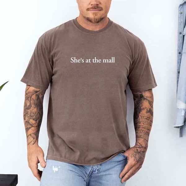 May include a man wearing an espresso Comfort Colors T-shirt with the phrase "She's at the mall" printed in white, styled casually with light denim jeans, showing tattooed arms in a relaxed pose.