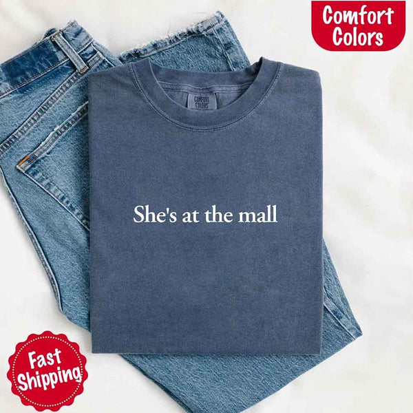 May include a folded denim Comfort Colors T-shirt with the text "She's at the mall" printed in white, styled with denim jeans and white sneakers, promoting fast shipping.