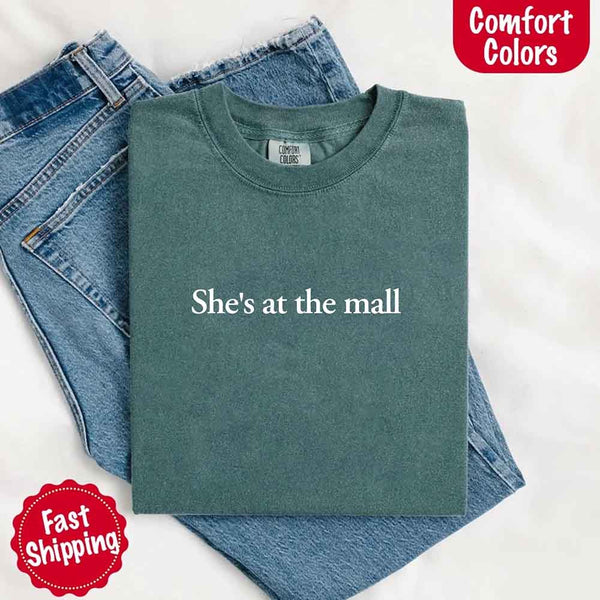 May include a blue spruce Comfort Colors T-shirt with the text "She's at the mall" printed in white, styled with denim jeans and white sneakers, promoting fast shipping.