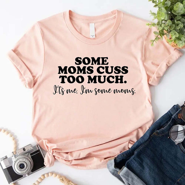 Peach t-shirt with humorous text some mom cuss too much on a white surface with accessories.