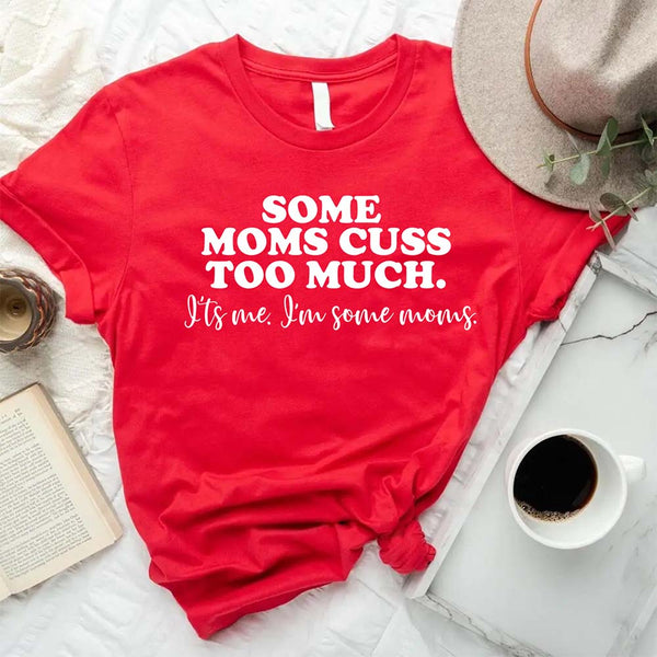 Red t-shirt with humorous text on some mom cuss too much  a white surface with a hat, book, and coffee cup.