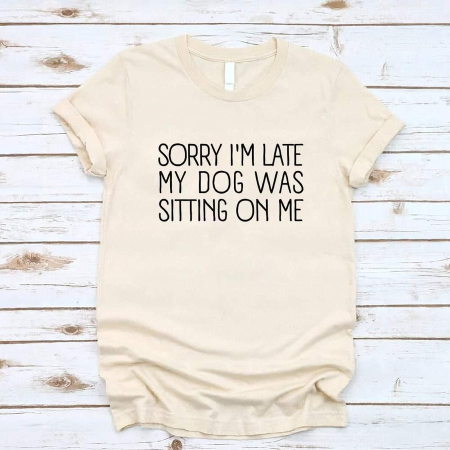 t-shirt with 'Sorry I'm late my dog was sitting on me' text, paired with jeans and sneakers on a wooden surface.