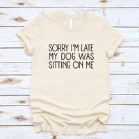 t-shirt with 'Sorry I'm late my dog was sitting on me' text, paired with jeans and sneakers on a wooden surface.