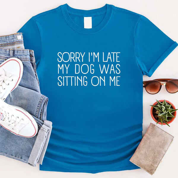 t-shirt with 'Sorry I'm late my dog was sitting on me' text, paired with jeans and sneakers on a wooden surface.