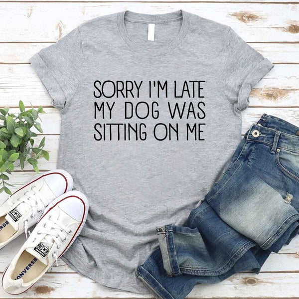 Gray t-shirt with 'Sorry I'm late, my dog was sitting on me' text, paired with jeans and sneakers on a wooden surface.