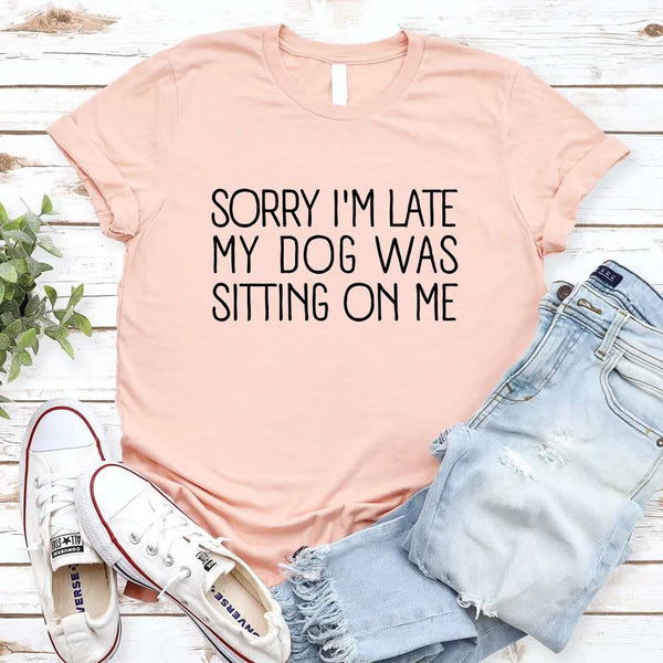 Peach t-shirt with text 'Sorry I'm late my dog was sitting on me' on a wooden surface with jeans and sneakers.