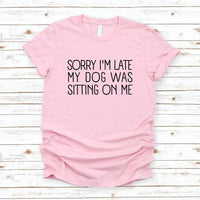 Pink t-shirt with text 'Sorry I'm late my dog was sitting on me' on a wooden surface