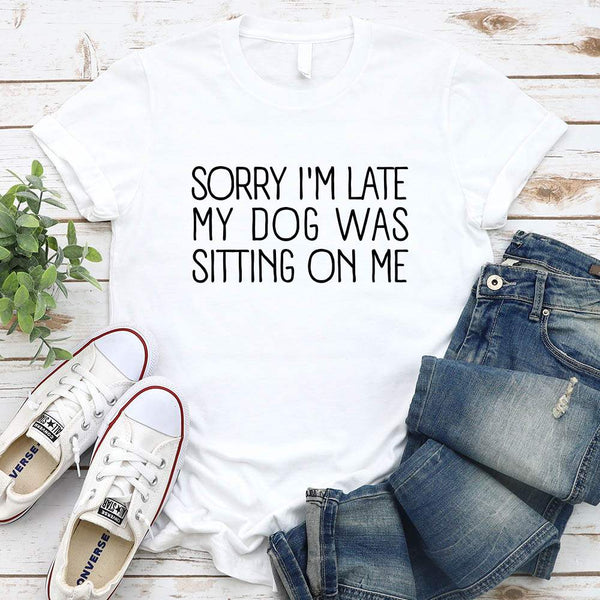 White t-shirt with text 'Sorry I'm late my dog was sitting on me', paired with jeans and sneakers on a wooden surface.