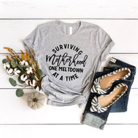 May include a white unisex graphic T-shirt laid flat with the words “Surviving Motherhood One Meltdown at a Time” printed in bold lettering across the chest.