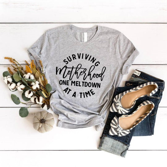 May include a white unisex graphic T-shirt laid flat with the words “Surviving Motherhood One Meltdown at a Time” printed in bold lettering across the chest.