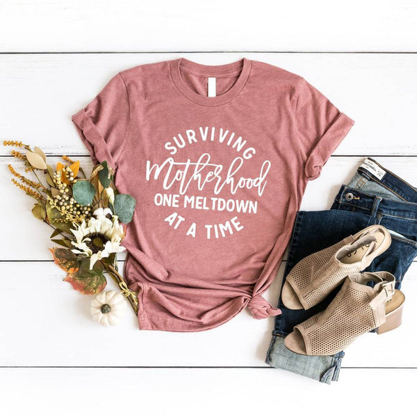 May include a white unisex graphic T-shirt laid flat with the words “Surviving Motherhood One Meltdown at a Time” printed in bold lettering across the chest.