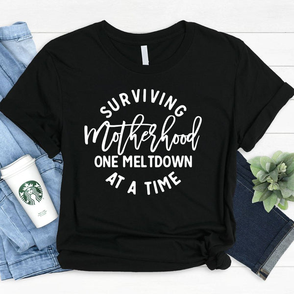 May include a white unisex graphic T-shirt laid flat with the words “Surviving Motherhood One Meltdown at a Time” printed in bold lettering across the chest.